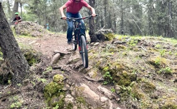 A youth confidently rides their bicycle down a rocky and rooty section of trail